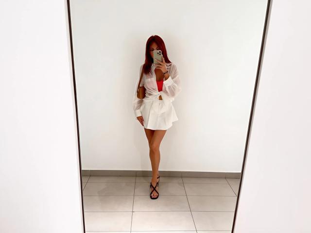 Photographie d'AlisaxLoveX debout dans une pièce très éclairée, portant une robe blanche avec un nœud rouge, tenant un smartphone. jambes légèrement écartées, sandales noires, contraste élevé, tension sexuelle.