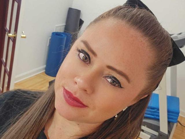 Foto em close-up de ChantalNoa, uma mulher de pele clara, cabelo castanho preso em um rabo de cavalo, faixa de cabeça preta, batom vermelho e roupa preta. delineador, olhando para a câmera com um leve sorriso. Ela está vestindo uma blusa preta. O fundo inclui uma bola de exercícios azul e equipamentos de ginástica. Muito atraente..