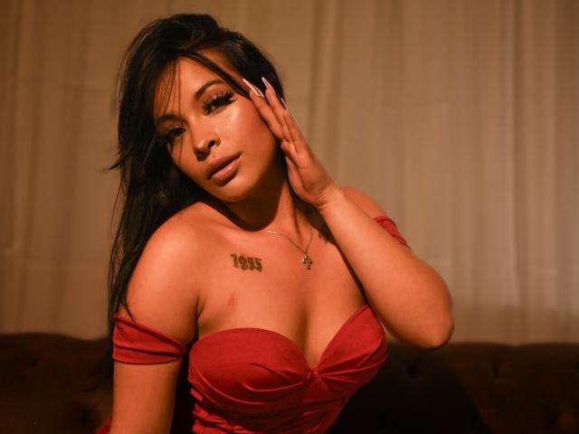 Fotografía de MiaMillerr69, una mujer de pelo oscuro y largo, que lleva un vestido rojo con un hombro descubierto, con una mano en la mejilla, con una expresión seductora. mirada, piel marrón claro, tatuaje en el pecho, poca luz, colores cálidos.