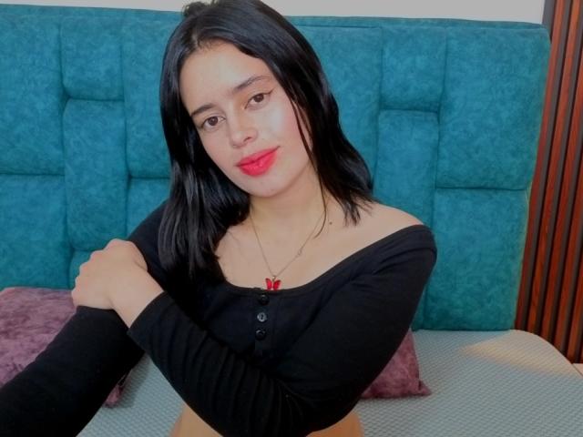 JuneCute, con i capelli neri e il rossetto rosso, è seduta su un letto con le braccia incrociate, indossando una maglietta nera a spalle scoperte e un paio di pantaloni rossi. collana con pendente, su uno sfondo di una testiera imbottita color verde acqua. Assolutamente attraente..