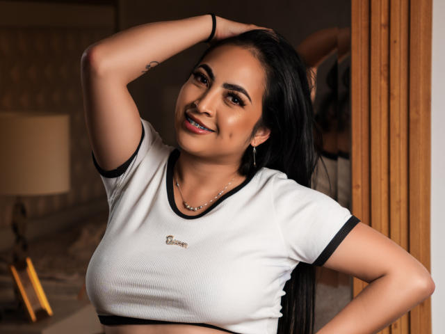 Photographie d'Alana West avec de longs cheveux noirs, souriante, la main derrière la tête, portant un t-shirt blanc à rayures avec des détails noirs. yeux marron, peau claire, tatouage discret sur l'avant-bras, pièce faiblement éclairée.