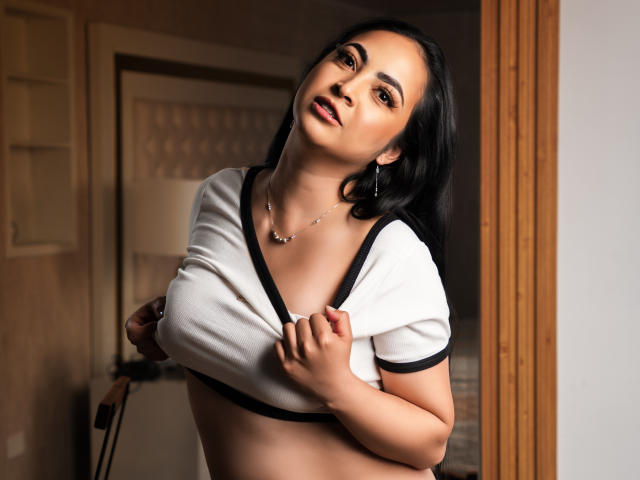 Fotografía de Alana West con cabello oscuro, tono de piel medio y busto grande. Ella está levantando su blusa blanca con detalles negros, mostrando su escote. Ella la mira de forma seductora, con una ligera sonrisa. El fondo incluye una puerta de madera y una lámpara..