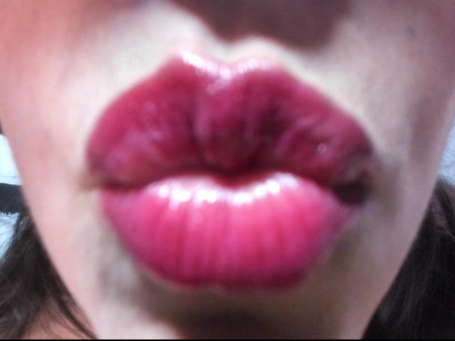 Fotografia ravvicinata del viso di DakotaGray, con particolare attenzione alle labbra lucide e di un rosso acceso, leggermente dischiuse, che rivelano un accenno di denti. L'illuminazione esalta la texture e il colore delle labbra, creando un effetto sensuale..