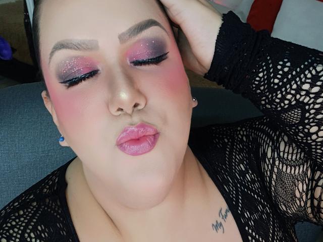 Fotografía de primer plano de GiiannaMorrys con colorete rosa, sombra de ojos púrpura brillante y labios rosados. Ella tiene una blusa de encaje negro, aretes azules y un tatuaje pequeño en el cuello. Sus ojos están medio cerrados de forma seductora..
