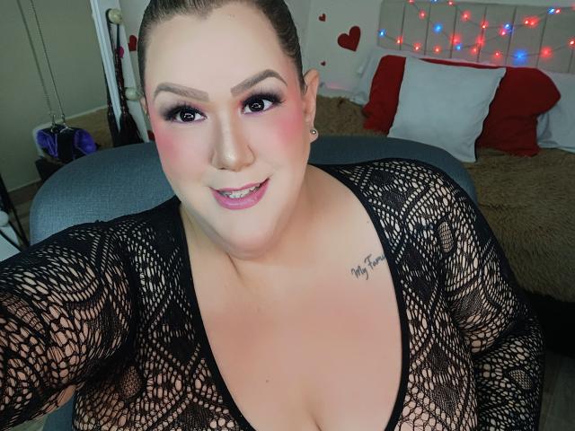 Nahaufnahme-Selfie von GiiannaMorrys, die lächelnd mit pinkem Lippenstift, dunklem Eyeliner und einem schwarzen Spitzenoberteil zu sehen ist, das das Dekolleté freilegt. Tätowierung an der rechten Schulter, rote und weiße Kissen im Hintergrund, Lichterketten. Verdammt heiß..
