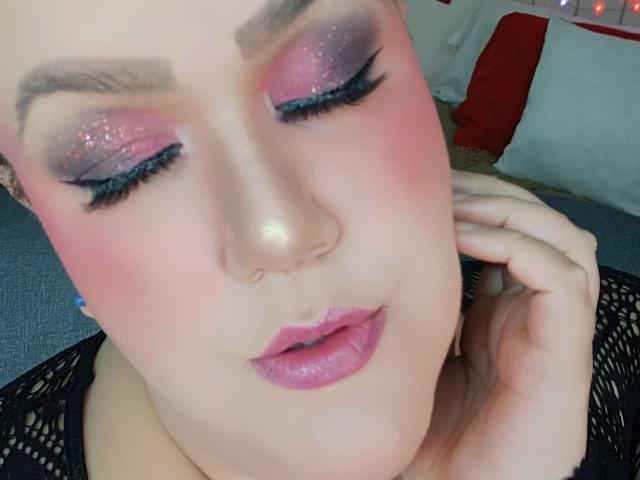 Nahaufnahme von GiiannaMorrys mit geschlossenen Augen, schimmerndem rosa Lidschatten, dicker schwarzer Eyeliner und rosa Lippenstift. Hand in der Nähe des Gesichts, schwarzes Spitzenoberteil sichtbar. Rouge betont das Gesicht, weiches Licht. Verführerischer Ausdruck.