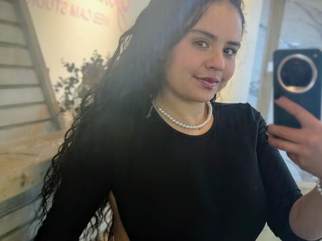ArielleWilde, a fair-skinned woman with long black hair, wearing a black dress and pearl necklace, takes a selfie. She has a slight smile, holding a smartphone with a circular ring.
