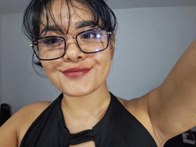 Close-up of JennLunaLee with messy black hair, wearing black halter top, glasses, and red lipstick, smiling slightly. Her arm is raised, revealing an armpit. Sexual tension, slight cock tease.