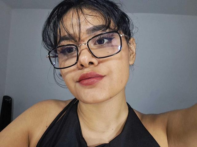 Close-up of JennLunaLee, a Latina woman with black hair in a messy bun, wearing black-framed glasses and a black halter top. She has red lipstick and a confident expression. Her gaze is direct.