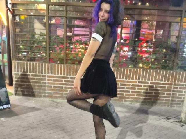 DarckVelvet è in piedi con una gamba sollevata, indossa una maglia nera trasparente, un bracciale bianco, una gonna nera e collant a rete. Capelli viola, sorriso sfrontato, sfondo cittadino illuminato al neon, mattoni e strada..