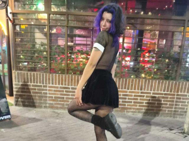 DarckVelvet, avec ses cheveux bleus, sa jupe noire et ses bas noirs transparents, se tient sur une jambe, regardant en arrière de manière séduisante. L'arrière-plan présente des lumières néon colorées et un mur en briques. Les doigts sont posés sur la hanche..