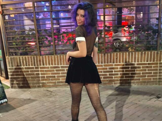 DarckVelvet, com cabelo roxo, está em uma rua pavimentada com tijolos, de costas, olhando por cima do ombro, vestindo uma saia preta e meias finas pretas. Luzes de neon refletem nas janelas de vidro ao fundo. Incrivelmente sexy..