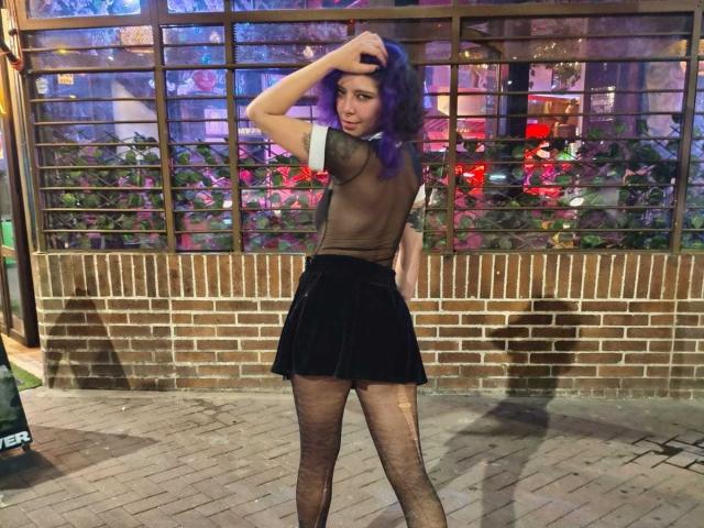 DarckVelvet, met paars haar, staat 's nachts op een straat, met de rug naar de camera, haar hand in haar haar, en draagt een zwarte, doorschijnende top, kort. een zwarte rok en netpanty. Neonlicht reflecteert achteraan..