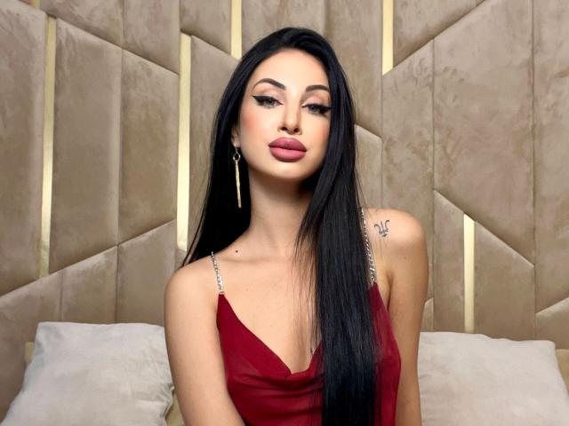CelineNoir, uma mulher atraente com longos cabelos pretos, vestindo um vestido vermelho com decote baixo, está sentada encostada em uma cabeceira bege com detalhes em botão. Seus lábios são cheios e pintados de vermelho, transmitindo confiança e atração..