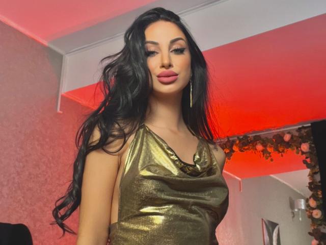 CelineNoir, een aantrekkelijke vrouw met lang zwart haar, gekleed in een glanzende gouden jurk, staat zelfverzekerd in een kamer met rode verlichting. Haar lippen zijn samengeknepen, wat een verleidelijke uitstraling geeft..