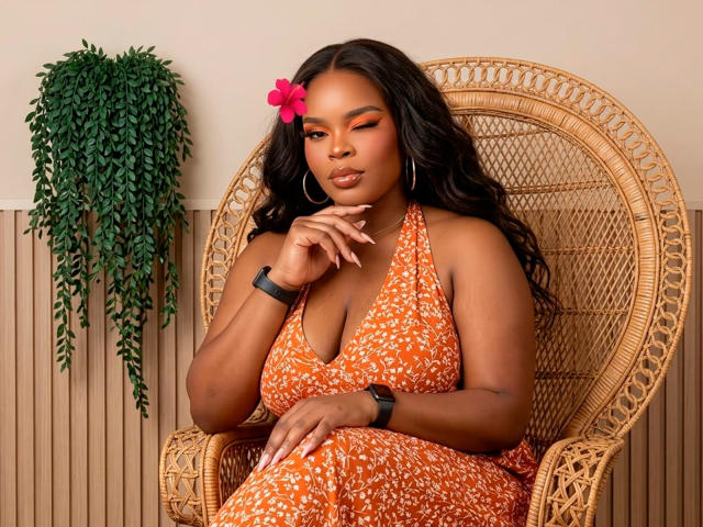 IsisGonzales sits in a wicker chair, wearing an orange floral dress with a deep neckline, highlighting her large breasts. She has long black hair with a pink flower, looking sultry.