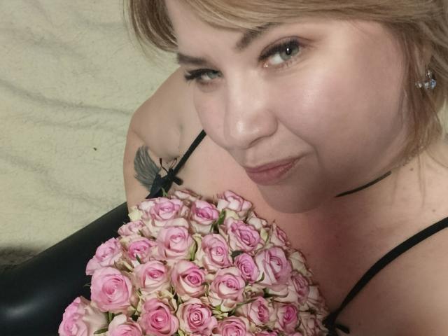 Close-up van CurlyDreams, een mollige vrouw met lichte huid en bruin haar, die een boeket roze rozen vasthoudt. Ze draagt een zwarte jurk met bandjes, en heeft een tatoeage op haar schouder. Ze heeft een lichte grijns..