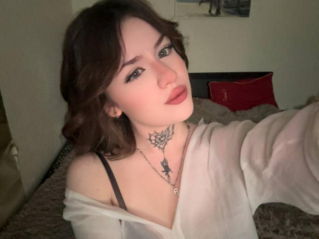 Een vrouw met een obsessie voor bleke huid, donkerbruin golvend haar en rode lippenstift, gekleed in een witte top met een open schouder en een zwarte band. zichtbaar, tatoeages op de nek, een selfie maken, verleidelijk in de camera kijken.