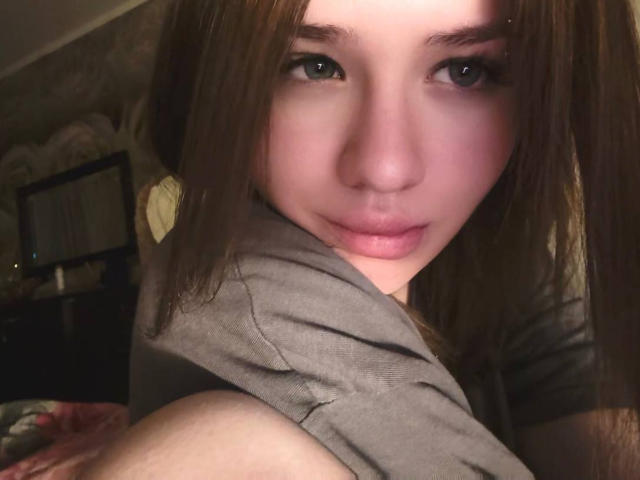 Gros plan de NeatObsession, avec une peau claire et des cheveux bruns foncés, regardant directement l'appareil photo avec des yeux intenses. Visage à moitié dans l'ombre, lumière douce sur la joue. Arrière-plan flou, sombre. Regard séduisant et intense.