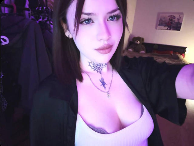 NeatObsession with pale skin, black hair, and blue eyes, wearing a low-cut white top, exposing cleavage, black tattoo on neck, black cardigan, bedroom setting, bed with stuffed animal, dim lighting.