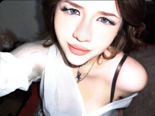 Close-up photo of NeatObsession with short brown hair, blue eyes, and a slight smile. She's wearing a white top with black straps, showing cleavage and a black necklace. Bright lighting.