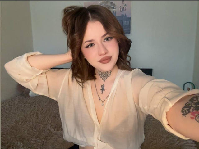 Selfie da NeatObsession, com pele clara e cabelo castanho ondulado, vestindo uma blusa branca transparente, colar preto com cruz, braço tatuagem, batom escuro, mão no cabelo, luz do sol realçando o rosto, pose casual, tensão sexual..