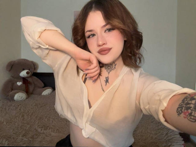 NeatObsession, con piel clara y cabello ondulado castaño, posa en un selfie, vistiendo una blusa blanca anudada que deja ver parte de su escote. con un collar negro y un tatuaje de estrella en el cuello. Un oso de peluche marrón está sentado en el fondo..