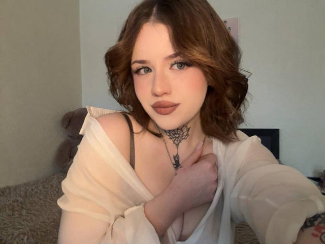 NeatObsession with fair skin, wavy brown hair, and dark lipstick, wearing a sheer white top revealing a black bra and lace choker. Hand gripping top, sexual tension, seductive gaze.