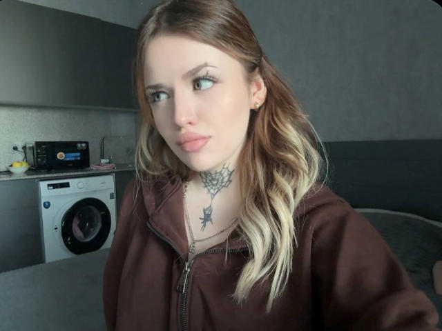 Une jeune femme, obsédée par la peau pâle et les longs cheveux blonds ondulés, portant un sweat à capuche marron, est assise devant une machine à laver. Elle a un tatouage noir sur sa poitrine. Un regard incroyablement sexy et intense..