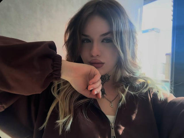 NeatObsession, una donna attraente con capelli biondi ondulati, indossa una giacca marrone scuro e si sporge in avanti, con la mano vicino alla bocca. la luce del sole che illumina il suo viso, sguardo intenso, collana visibile, sensualità palpabile..
