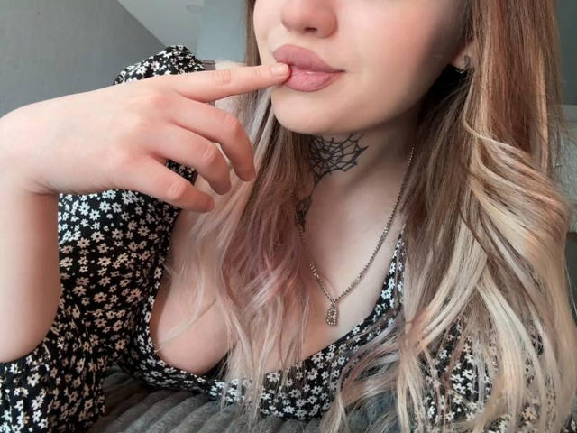 Close-up photo of NeatObsession, fair-skinned with blonde ombre hair, biting her finger while wearing a black floral dress. Visible chest tattoo, necklace. Sexy, seductive expression, slightly parted lips.