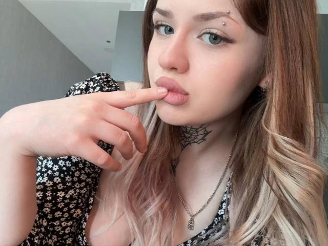 NeatObsession tem pele clara e cabelo castanho com pontas loiras, lábios carnudos e um dedo que a toca de forma sedutora. Ela está vestindo um vestido floral preto, um colar e um colar de renda preto..