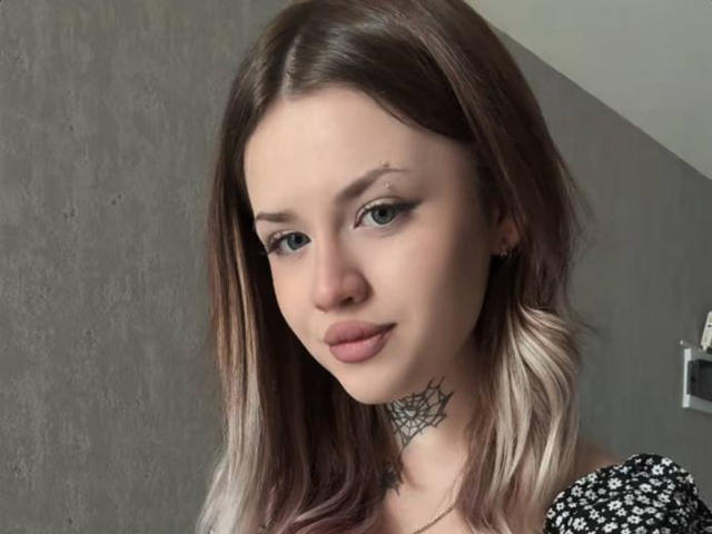 NeatObsession has shoulder-length brown hair with blonde highlights, wearing a black and white patterned top. She has a spiderweb tattoo on her neck, looking at the camera with a slight smile.