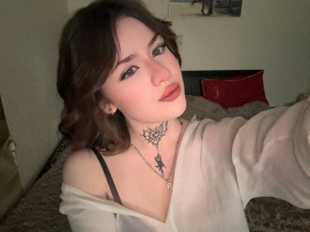 NeatObsession, met een bleke huid en donkerbruin haar, draagt een witte top met een open schouder, met een zwarte band zichtbaar, en heeft tatoeages op nek, lippen lichtjes open, donkere oogmake-up, voorovergebogen, verleidelijke blik.