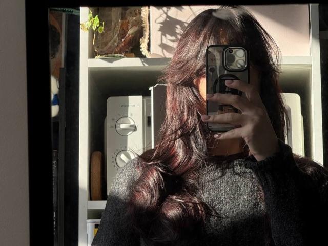 Tsukerberg mit langem, gewelltem, braunem Haar, der ein Selfie mit einem Smartphone macht, sein Gesicht im Schatten, ein schwarzer Pullover, hell. Sonnenlicht wirft Schatten, ein Regal mit Pflanzen und Gegenständen im Hintergrund..