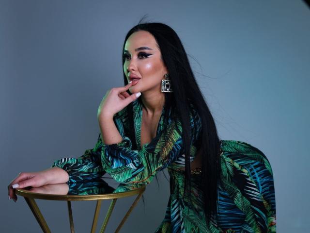 XandraRaven leans seductively over a gold table, wearing a green and blue patterned dress. She has long black hair, large earrings, and a sultry expression with her finger on her lips.