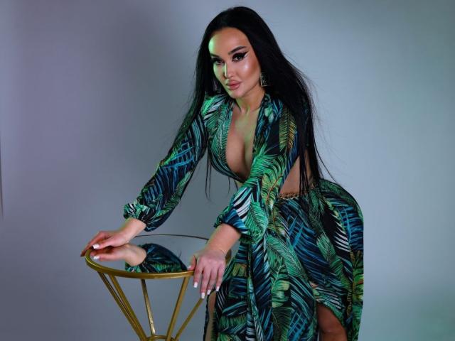 XandraRaven, una donna formosa con lunghi capelli neri, si sporge in avanti su una sedia dorata, indossando un abito verde e blu con motivi, abito con scollatura profonda, che mette in mostra un generoso décolleté e una spaccatura alta fino alla coscia. L'illuminazione al neon mette in risalto la sua espressione sensuale..