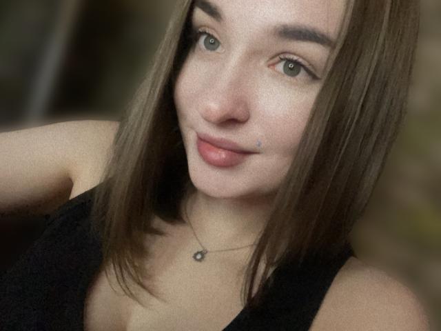Nahaufnahme von Lilit69, einer jungen Frau mit heller Haut, braunen, schulterlangen Haaren und blauen Augen, die ein schwarzes Tanktop trägt. Sie hat einen kleinen Schönheitsfleck unter ihrem linken Auge und eine silberne Halskette. Verdammt heiß, leichtes Lächeln, direkter Blick..