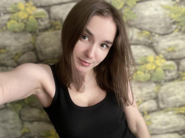 Lilit69 se toma un selfie con una ligera inclinación, cabello castaño, piel clara y una camiseta negra sin mangas. Está de pie frente a una pared de piedra desgastada, con manchas de musgo amarillo. Qué lindo/a..