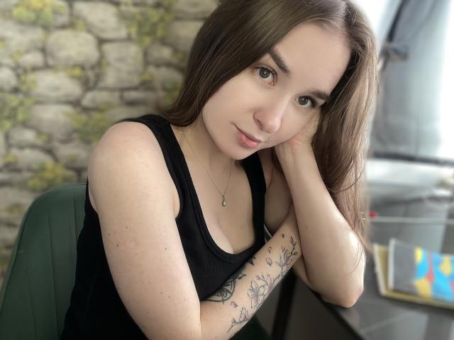 Lilit69, una joven de piel pálida y cabello largo y castaño, se apoya en su mano, vistiendo una camiseta de tirantes negra y con un brazo tatuado. tensión sexual evidente, mirada seductora, iluminación suave, fondo desenfocado.