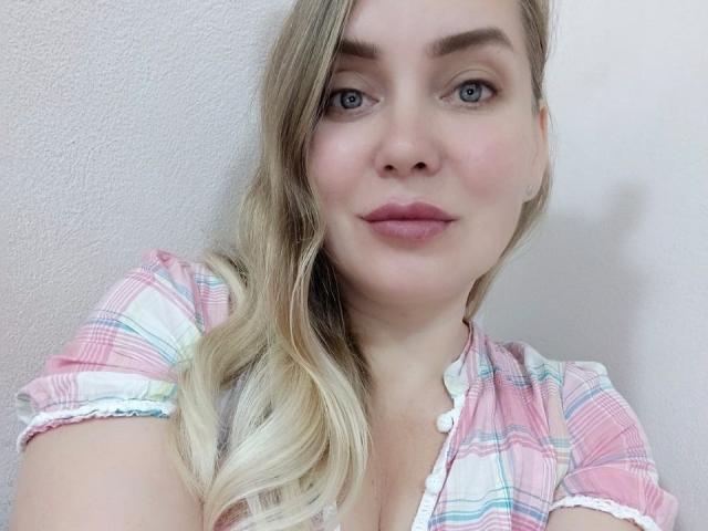 Close-up de LolaWhiter, uma mulher loira com olhos azuis e lábios rosados, vestindo uma camisa xadrez rosa. Seus cabelos ondulados caem sobre o ombro. Ela olha diretamente para a câmera com um sorriso discreto..