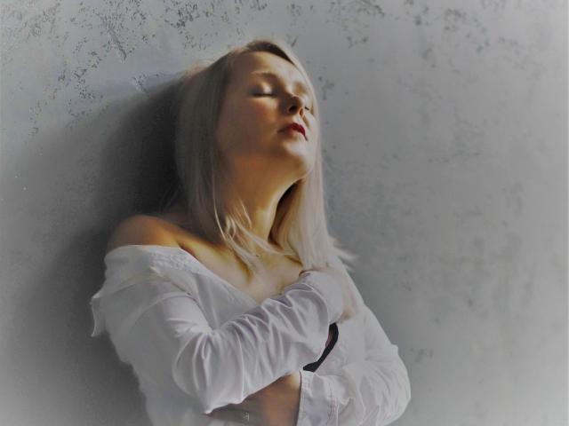 Fotografia di LolaWhiter con la pelle chiara e i capelli biondi, appoggiata a un muro strutturato, con gli occhi chiusi e la testa reclinata all'indietro. indossando una maglietta bianca con una spalla scoperta, che emanava una forte tensione sessuale.
