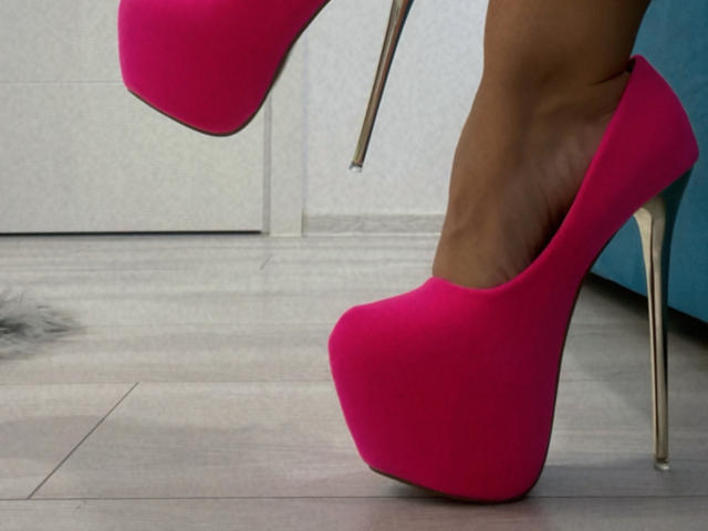 Primo piano del piede di MoniqueBliss, che indossa un paio di décolleté con tacchi a spillo alti e di un vivace colore rosa, con tacchi argentati. La sua pelle abbronzata contrasta con il tessuto rosa acceso. La tensione sessuale è suggerita dalla posa provocante..