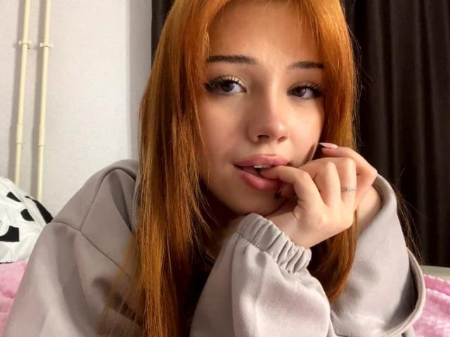 Close-up of SherrieDudycha with long red hair, light skin, and full lips, biting her finger. She wears a grey hoodie, looking directly at the camera with a sultry expression.