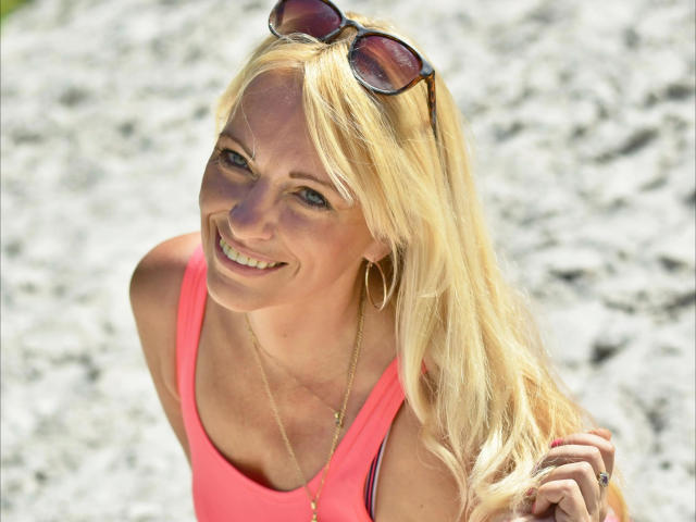Femme blonde, SofieHot-sex, souriant vers la caméra, portant un débardeur rose néon, des lunettes de soleil sur la tête, en or. collier, éclairé par le soleil, arrière-plan flou, lumineux, décontracté, sexy, ambiance estivale..