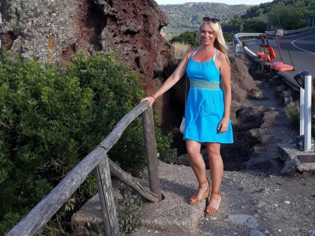 Sofie: Sexo apasionado, mujer rubia con un vestido azul brillante, apoyada en una barandilla de madera rústica, camino rocoso, arbustos verdes, cuerpo curvilíneo. pechos grandes, sandalias informales, sexy, entorno al aire libre, carretera de montaña en el fondo..
