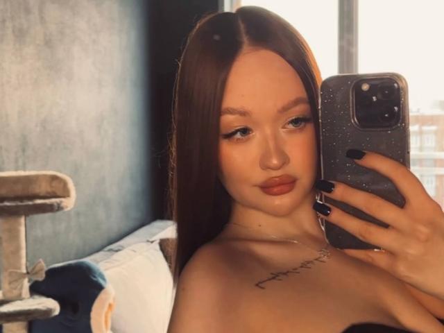 Foto van PamulaMetzner, onthuld, die een telefoon vasthoudt met zwarte nagels en een spiegel-selfie maakt. Lang bruin haar, rode lippen, kleine tatoeage op de linker schouder, zonlicht op de achtergrond, kattenkooi links..