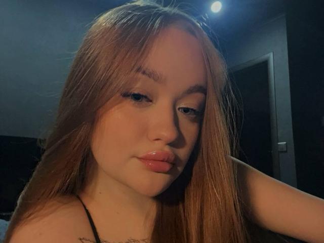 Gros plan de PamulaMetzner, une jeune femme aux longs cheveux bruns lisses, aux lèvres roses et au léger sourire. Elle porte un haut noir à bretelles, sous un éclairage bleu tamisé. Incroyablement sexy..