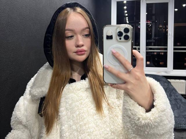 Foto van Pamula Metzner, een jonge vrouw met lang bruin haar, die een zwarte capuchon en een witte, pluizige jas draagt, en die iets vasthoudt. Witte iPhone met een popsocket, die een selfie maakt, aantrekkelijk, stedelijke omgeving..