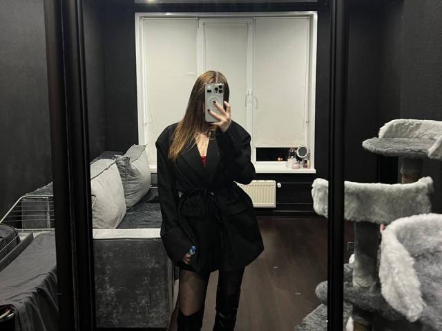 Fotografía de Pamula Metzner tomándose un selfie en un espejo, vistiendo un blazer negro y pantalones cortos negros, de pie en una habitación oscura. Con un rascador para gatos, una cama y una ventana iluminada al fondo. Increíblemente atractivo/a..